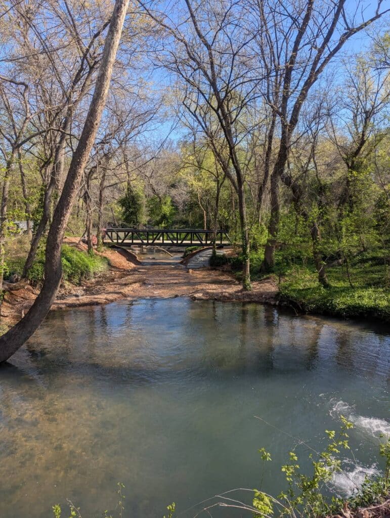 Peaceful creek flowing through a wooded area with a metal pedestrian bridge in the background.