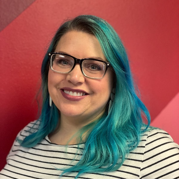 Smiling woman with long turquoise-blue hair and glasses wearing a striped shirt against a red background.