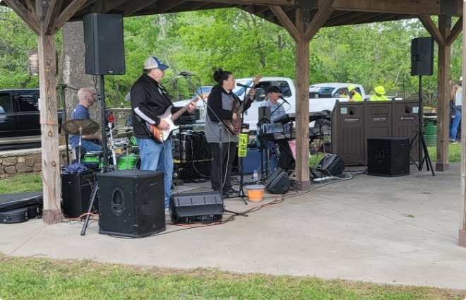Live band performing under an outdoor wooden pavilion, with musicians playing guitar, bass, and drums.