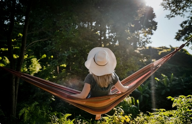 Person wearing a wide-brimmed hat relaxing in a colorful hammock surrounded by lush greenery and sunlight.