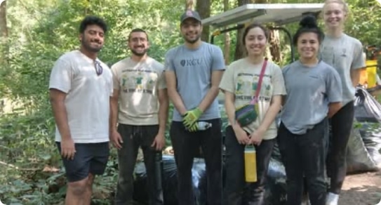 Six volunteers standing together outdoors in a wooded area after a cleanup activity, with collected trash bags behind them.