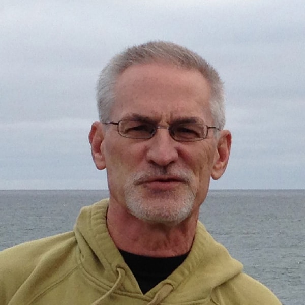 Man with short gray hair, glasses and a beard wearing a light green hoodie with a body of water in the background.