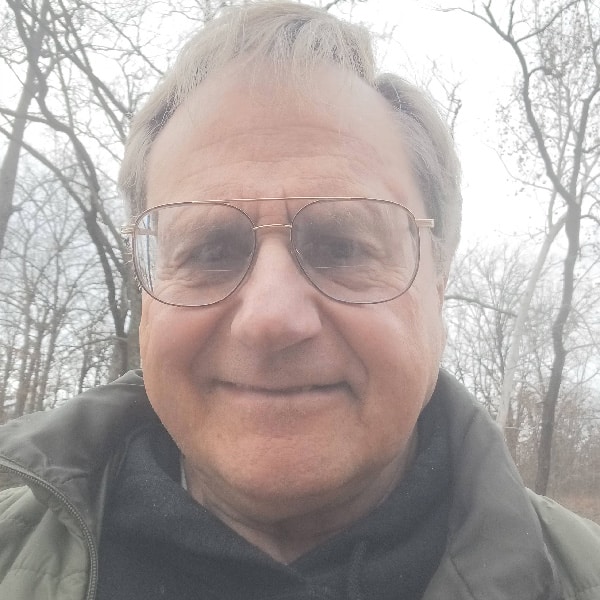 Man wearing glasses and a dark jacket smiling outdoors with trees in the background.