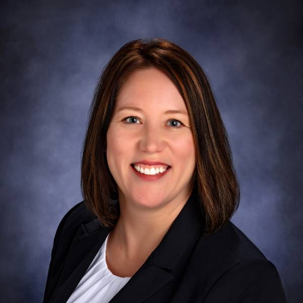 Professional headshot of a smiling woman wearing a dark blazer against a blue-gray background.